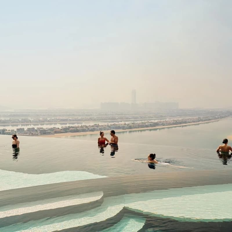 Rooftop infinity pool in Dubai with guests in the water, skyline haze, and panoramic hotel-view setting.