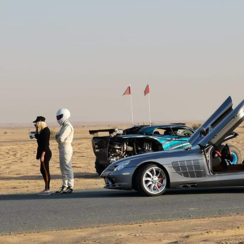 Desert automotive setup in Dubai with a silver supercar, off-road vehicle, crew, and open desert road environment.