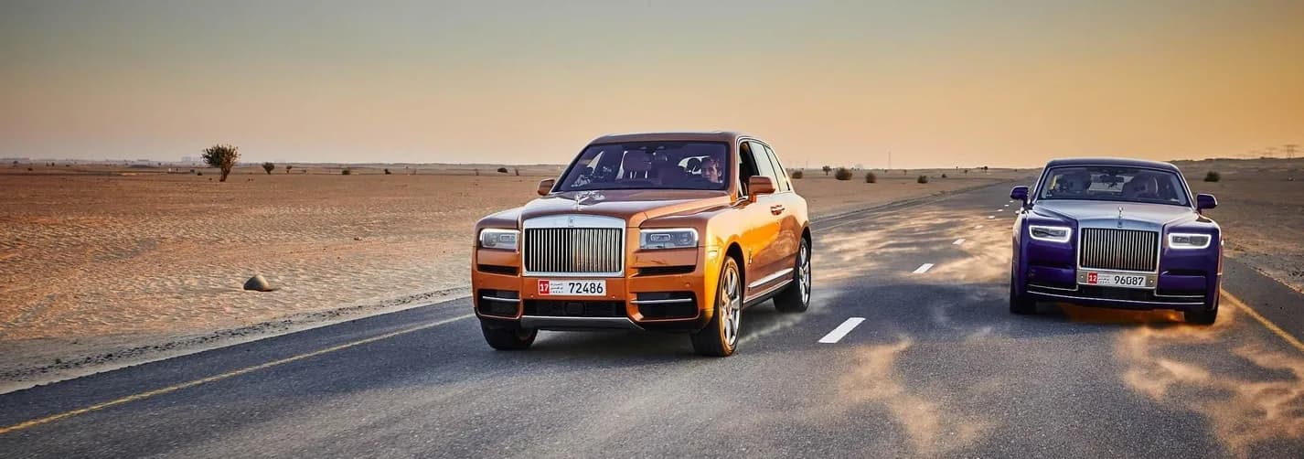 Two luxury vehicles driving on a desert road in Dubai with sand blowing across the asphalt at sunset.